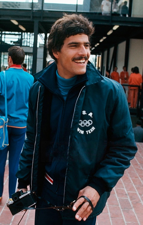 Competitive Olympic swimmer Mark Spitz wearing a forest green USA Olympic Swim Team Jacket holding a camera